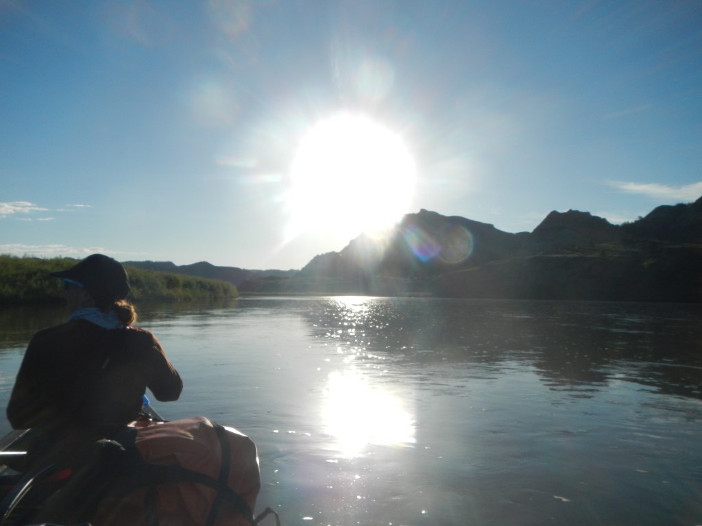 A person in a canoe floats down the river toward the sun. Mountains can be seen in the distance.